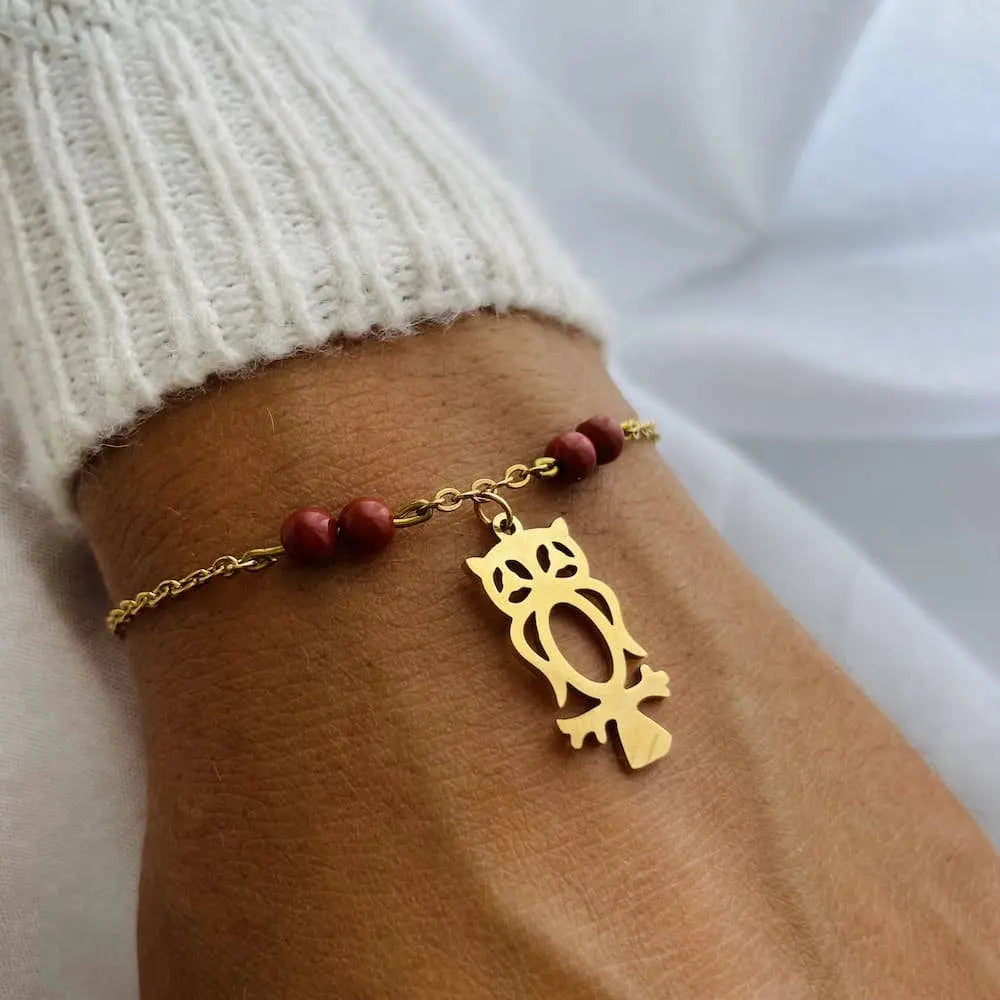 Elégant bracelet charm avec pendentif hibou doré et pierres rouges au poignet, symbolisant la sagesse et l'intuition en lithothérapie.